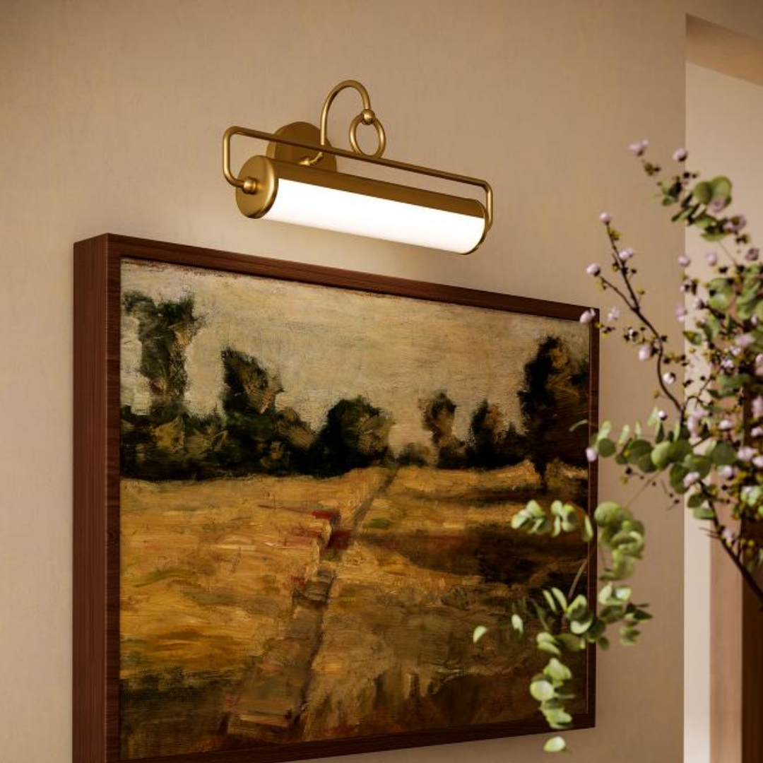 Brass LED picture light mounted above a framed landscape painting, illuminating artwork in a warm-toned hallway with wooden cabinetry and floral decor.