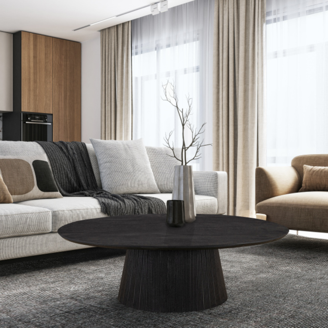 Modern living room with a round black coffee table, neutral-toned sofas, textured throw pillows, and floor-to-ceiling beige curtains in natural light.
