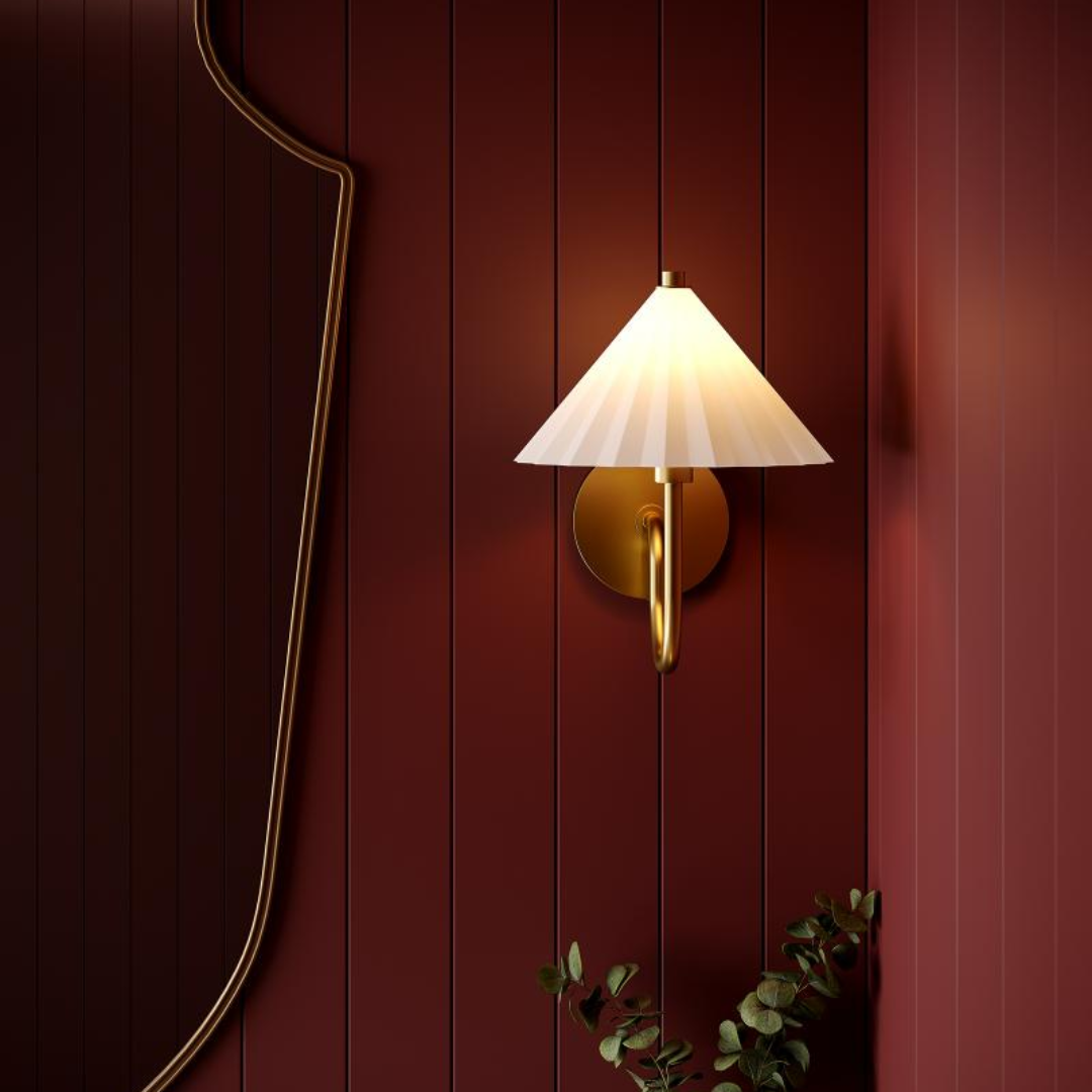 Elegant brass wall sconce with pleated white shade mounted on a deep red paneled wall, styled with a decorative mirror and greenery.